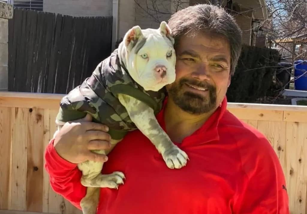 A man in a red shirt holding a white bulldog wearing a camouflage jacket.