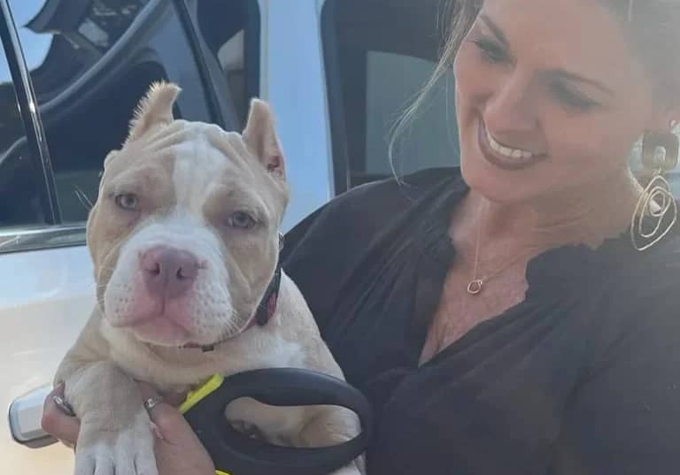 A woman holding a light brown puppy with a black collar near a car.